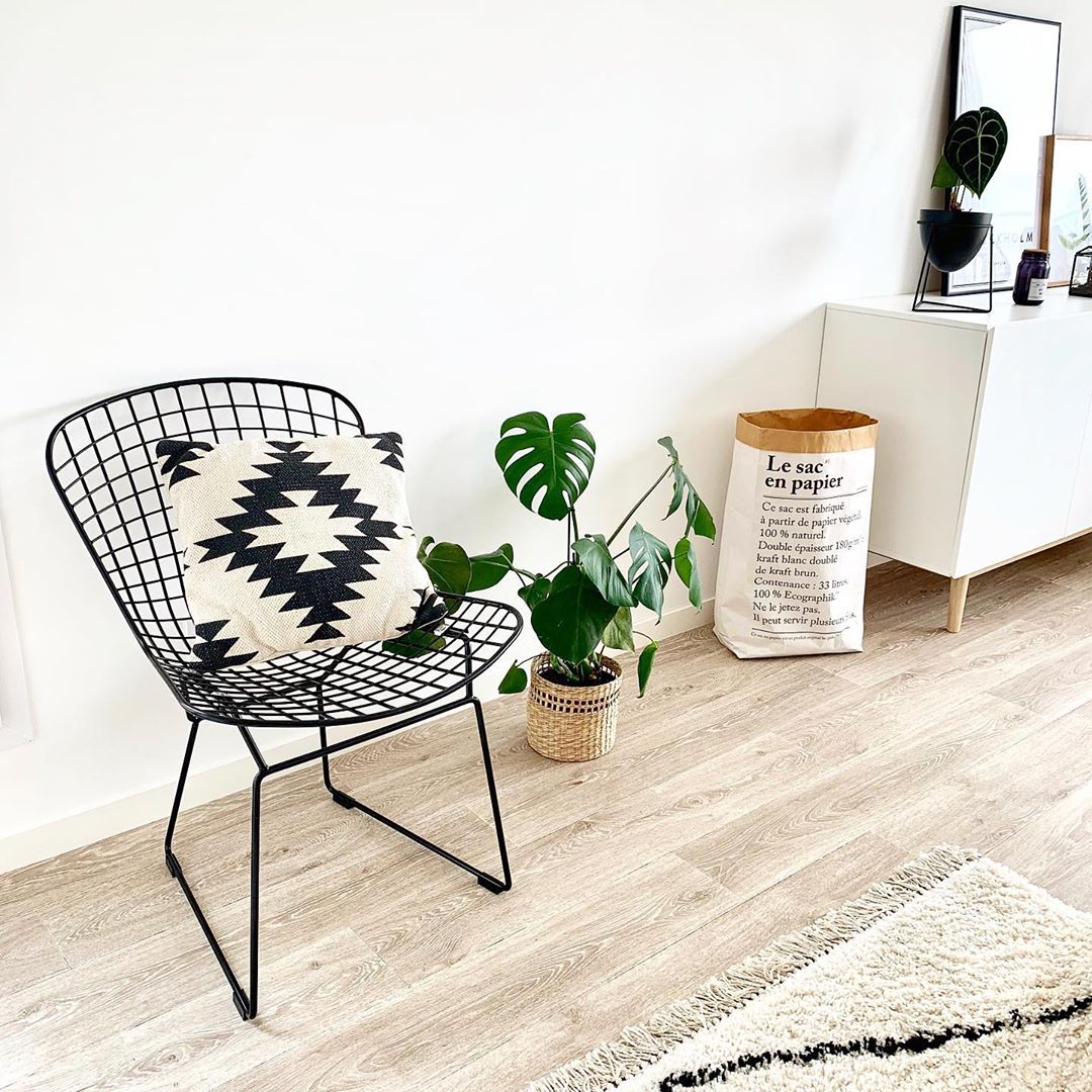 Chaise Diamant en m&eacute;tal de couleur noire dans une piece blanche avec un parquet en bois beige sur lequel se trouve un tapis laineux beige et noir avec des franges. Sur la chaise se trouve un coussin beige et noir &eacute;galement. A droite de cette chaise se trouve une plante, un sac en papier et une commode blanche avec des pieds en bois clair. Sur cette commode repose une plante dans un pot noir et des cadres. 